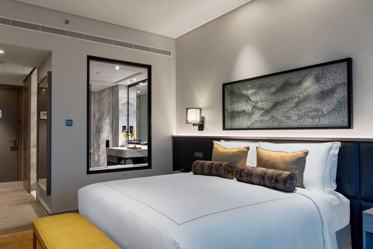 Habitació d'hotel moderna amb un llit gran, una taula de nit, una banca, un mirall i una obra d'art penjada a la paret.