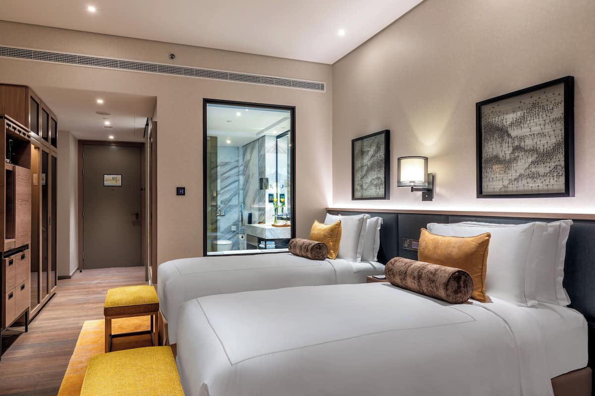 Habitació d'hotel amb dos llits, un armari de fusta, un bany visible a través d'una porta de vidre i obres d'art a les parets.