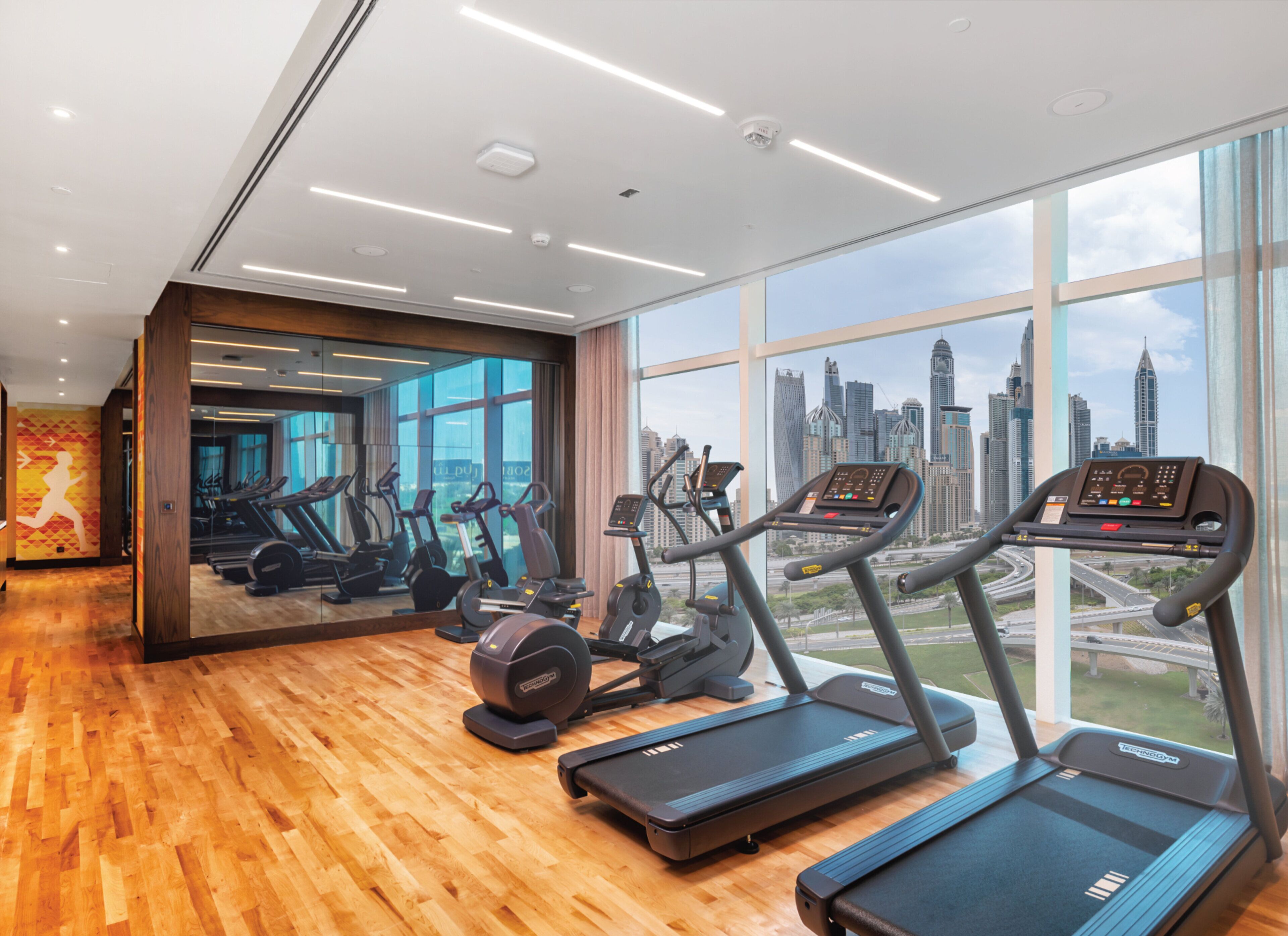 A gym with treadmills and elliptical machines, large windows with a city skyline view, and wooden flooring.