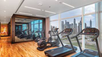 A gym with treadmills and elliptical machines, large windows with a city skyline view, and wooden flooring.