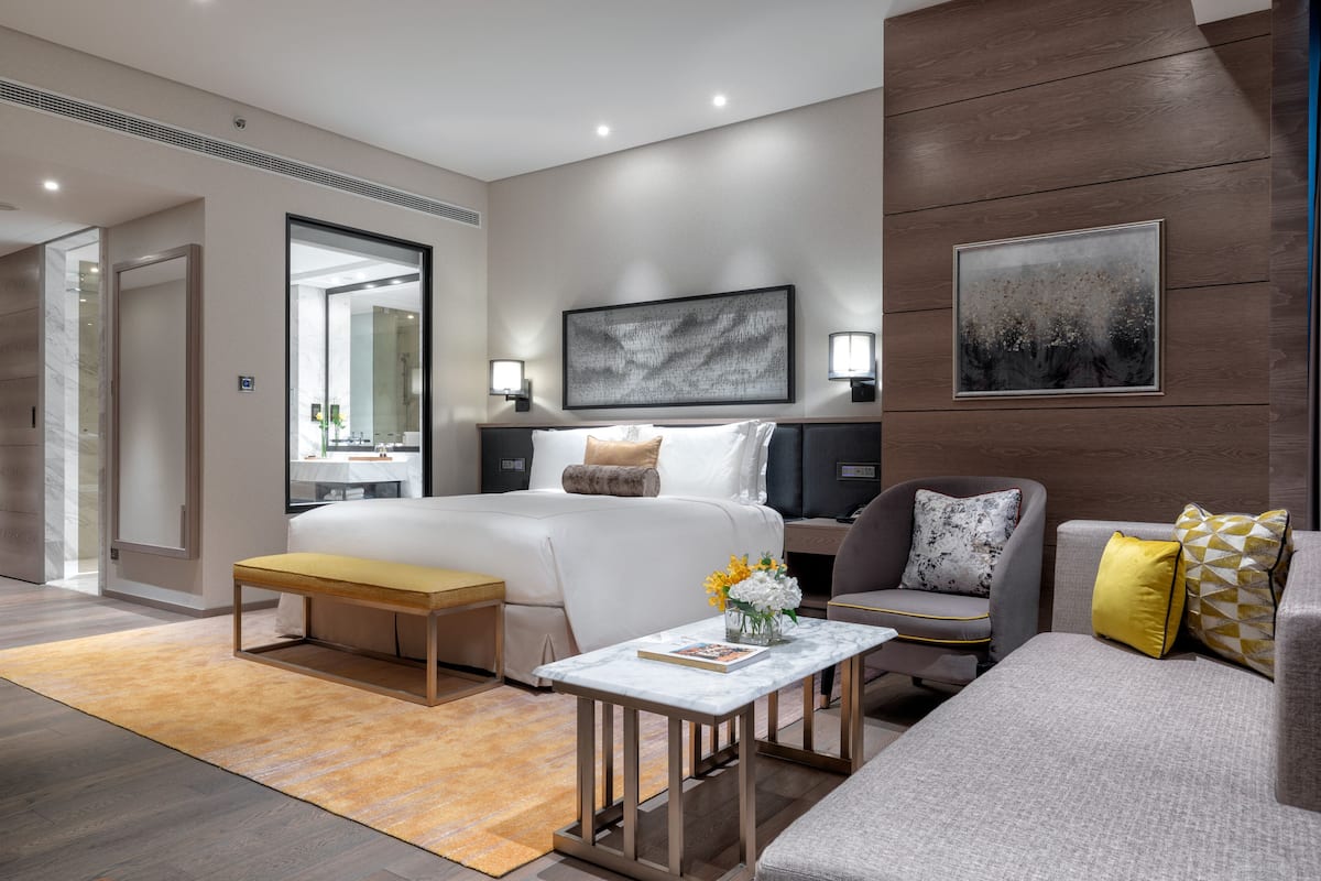 Una habitació d'hotel moderna amb un llit, un sofà, una taula petita i una cadira. Hi ha un mirall gran, un quadre a la paret i una finestra amb cortines.