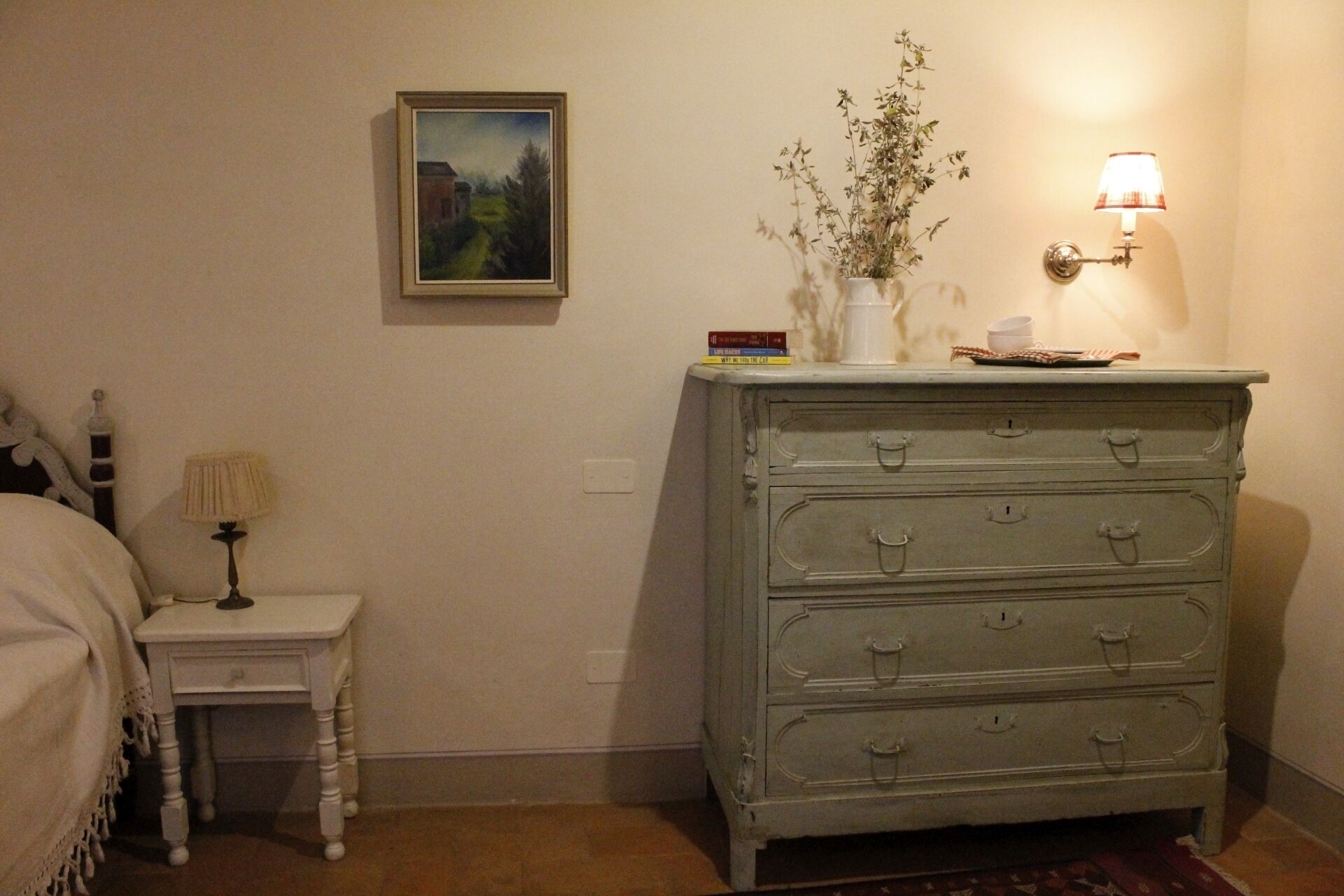 A bedroom with a bed, a nightstand, a dresser, a wall-mounted lamp, and a framed painting.
