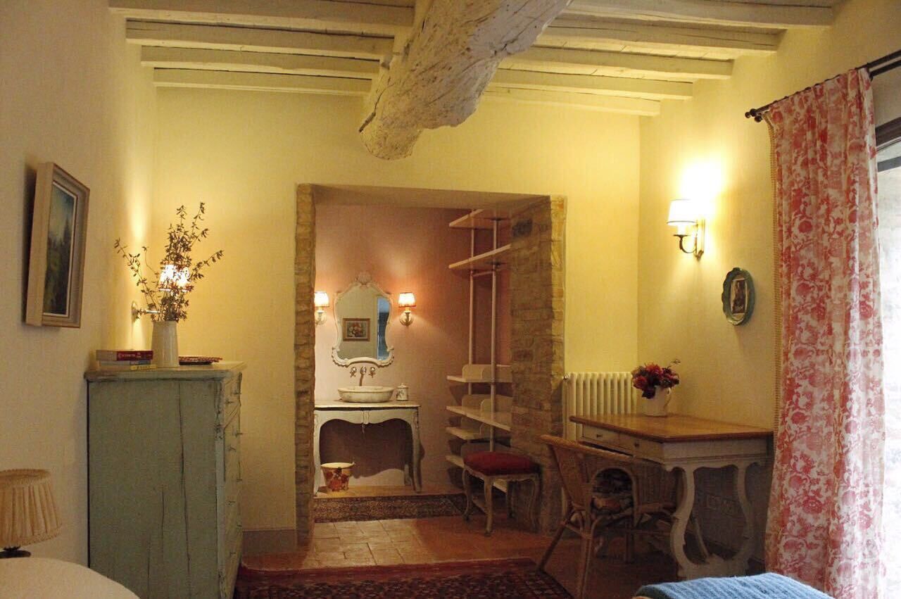 Una habitación con pared de piedra, un escritorio de madera, una silla, una mesita con un florero y una cama.