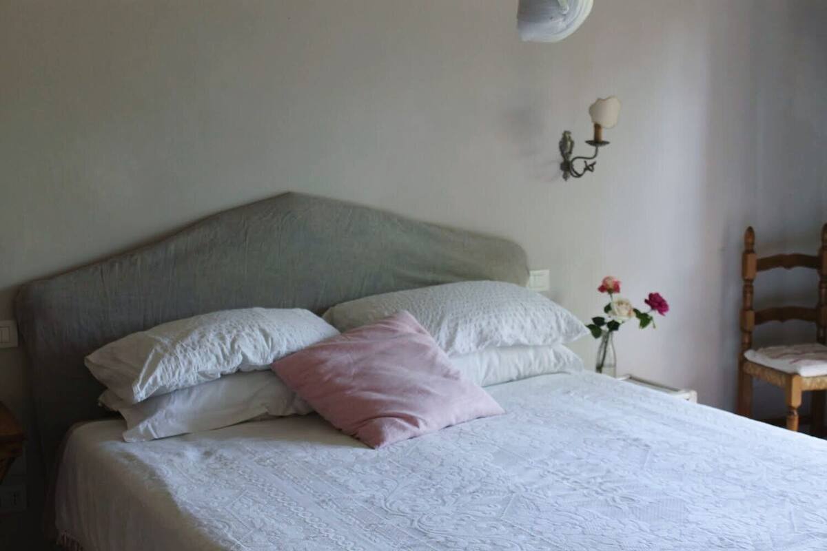 A bedroom with a bed, pillows, a nightstand, a vase with flowers, and a wall-mounted light fixture.