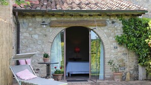 A stone building with a tiled roof, an open doorway, a bed inside, a lounge chair outside, and potted plants. - Private Room in small medieval borgo (Colle di Val d'Elsa)