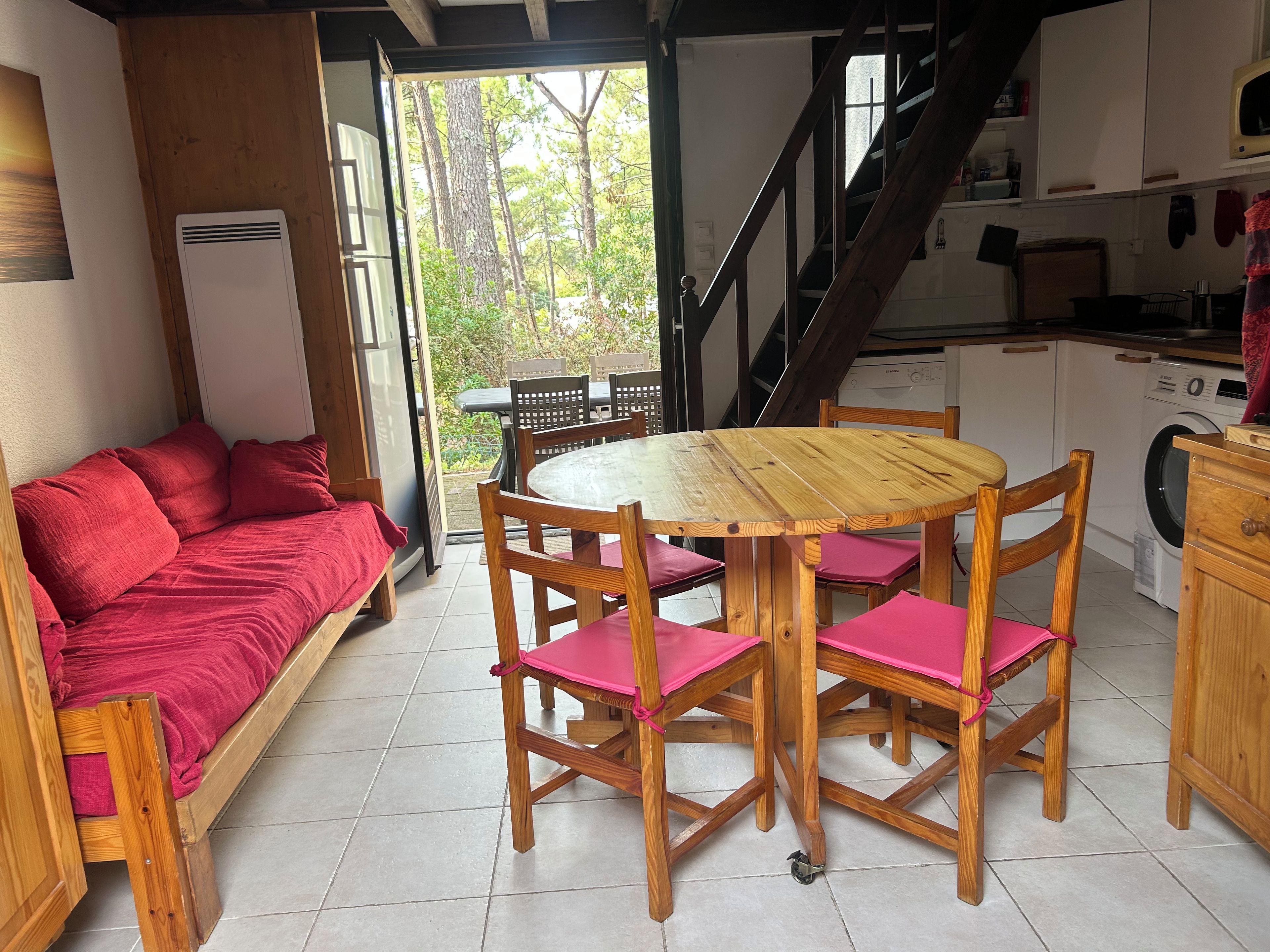 Une pièce chaleureuse avec un canapé en bois, une table ronde en bois entourée de chaises, un coin cuisine équipé d’un lave-linge et un escalier menant à un niveau supérieur.