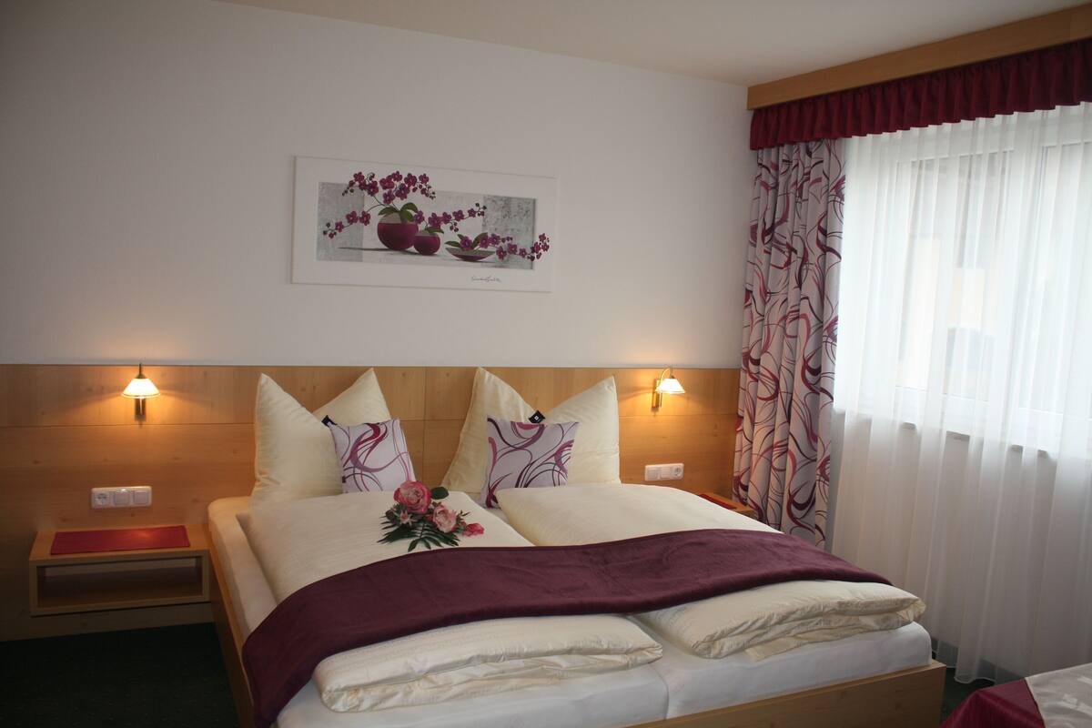 Una cama bien hecha con un arreglo floral, dos lámparas de noche y una obra de arte enmarcada sobre la cama.