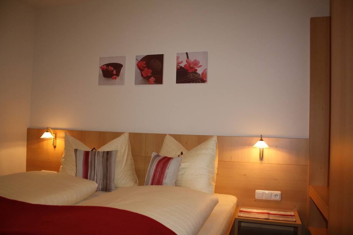 Habitación de hotel con cabecera de madera, dos lámparas de noche, una cama con manta roja y tres cuadros florales enmarcados en la pared.