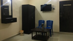 A small room with a black wardrobe, a TV mounted on the wall, two blue chairs, a small table, and a trash bin. - Hotel Duke Inn (Jhansi)