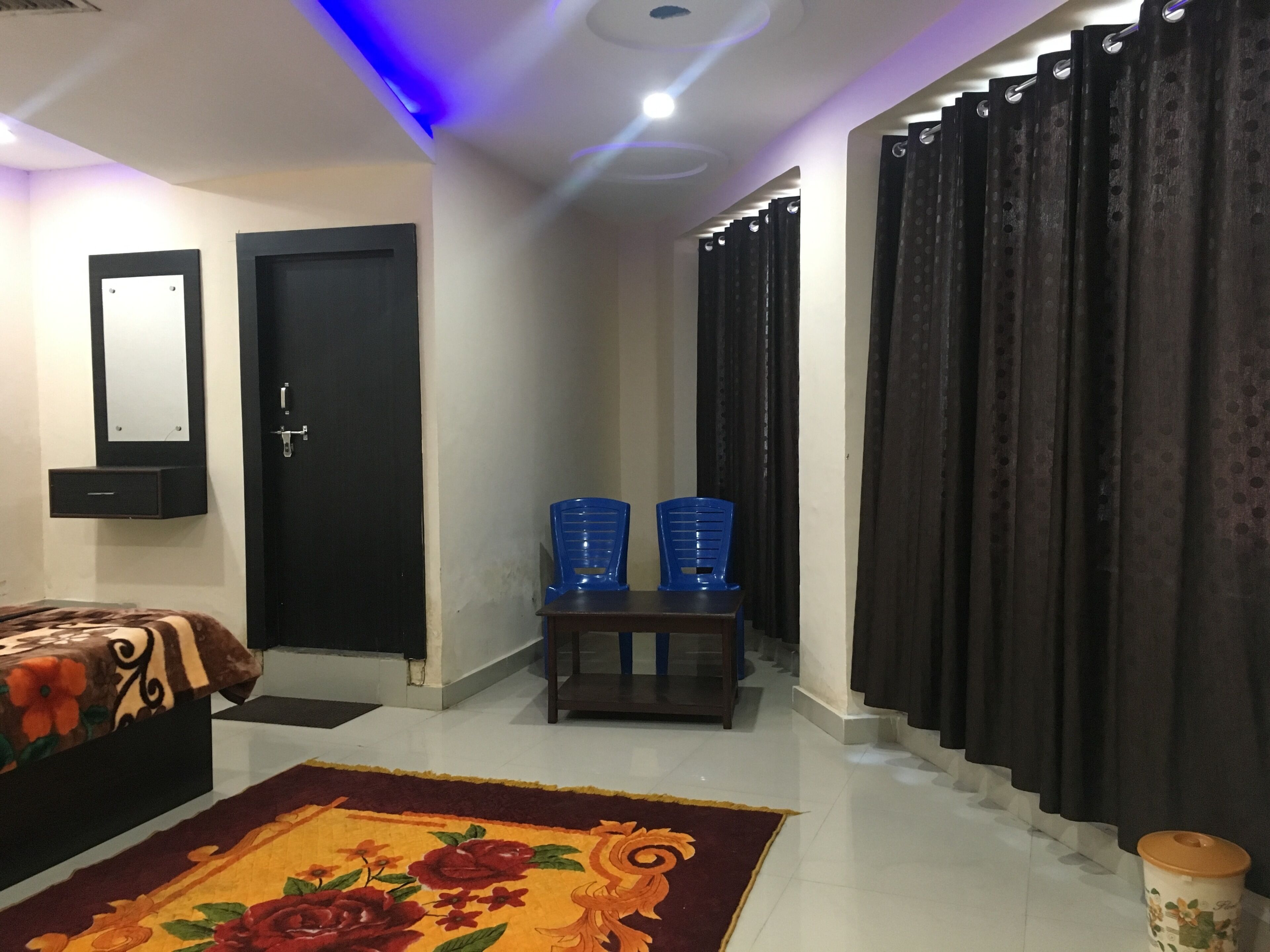 Une chambre d’hôtel avec un lit, une petite table, deux chaises bleues, une porte noire et un rideau.