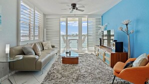 Una sala de estar moderna con un sofá, una mesa de centro y un sillón naranja. Hay un ventanal con persianas, un ventilador de techo y vistas al exterior.