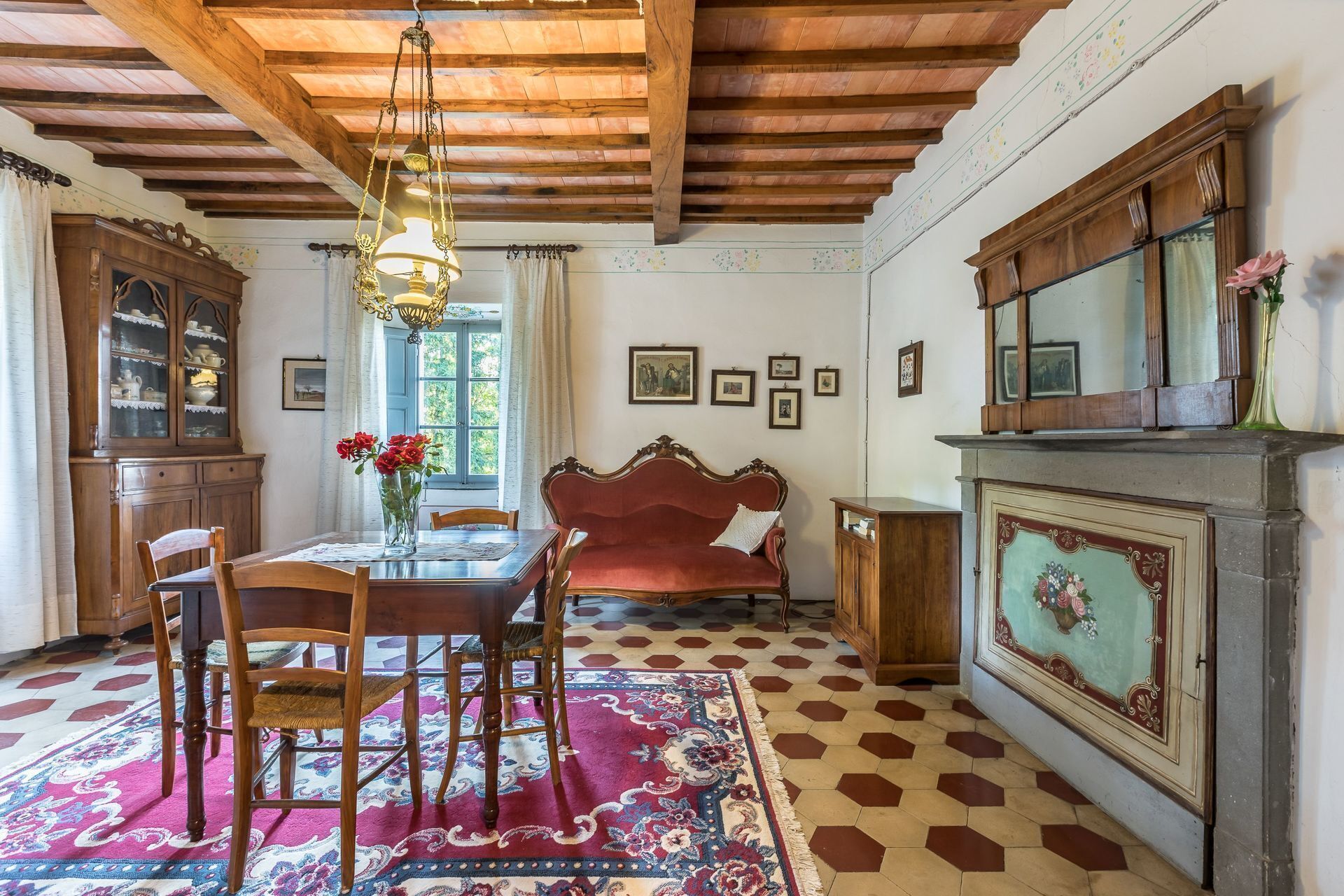 A dining area with a wooden table, chairs, a red sofa, a patterned rug, and a fireplace with a decorative panel.