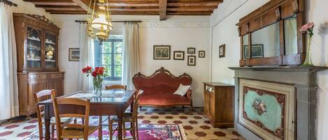 A dining area with a wooden table, chairs, a red sofa, a patterned rug, and a fireplace with a decorative panel.