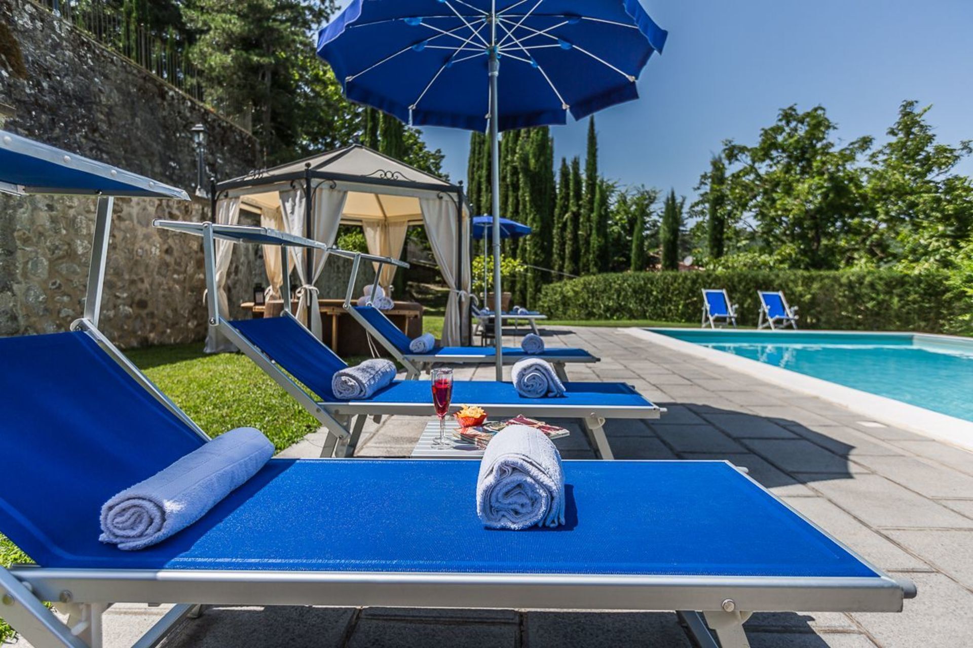 A poolside area with lounge chairs, a gazebo, and a blue umbrella.