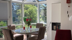 A conservatory with a glass table surrounded by chairs, a red chair, and a bookshelf with books.