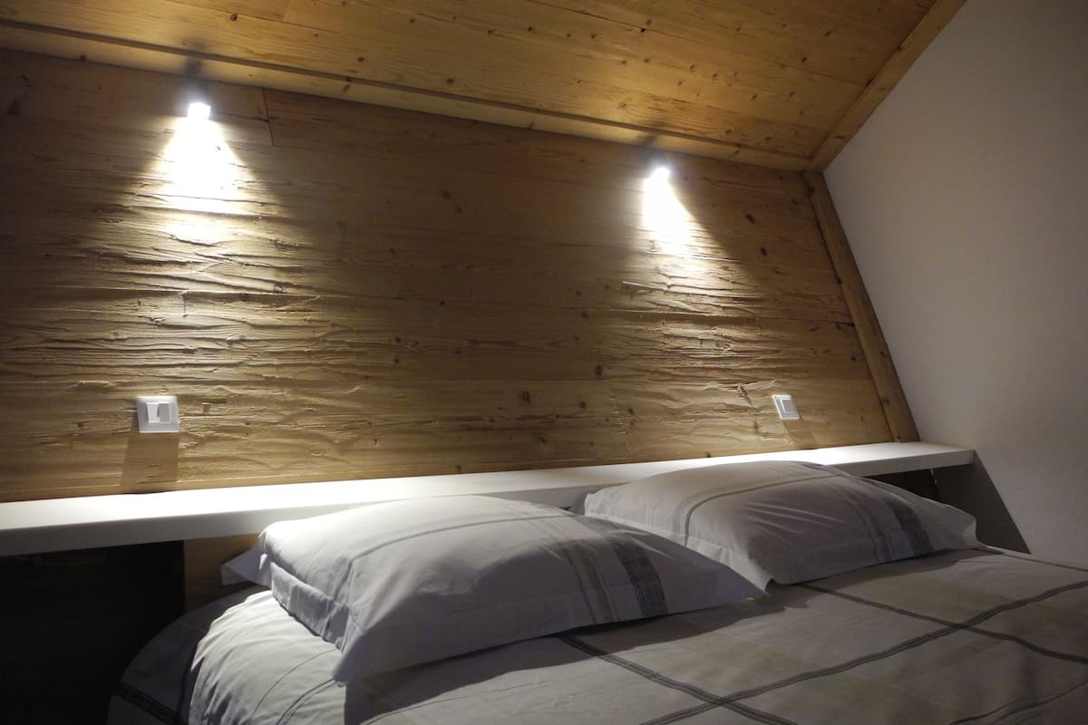Een slaapkamer met een houten plafond, twee bedlampen, een bed met een wit-grijs geruit beddegoed en twee kussens.