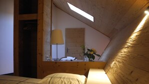 Un dormitorio con techo inclinado, vigas de madera, cama con ropa de cama blanca, mesita de noche con lámpara y ventana con luz natural.