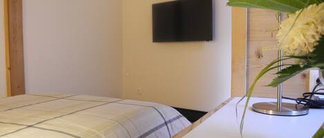A bedroom with a bed, a wall-mounted TV, and a bedside table with a lamp and flowers.