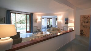 A spacious living area with a sofa set, a coffee table, and a dining table. There are large windows providing natural light and a view of the outdoors.
