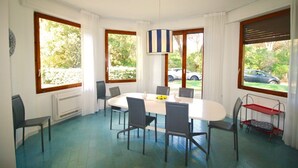 A dining room with a round table, six chairs, and a hanging light fixture. There are large windows with curtains, a radiator, and a small red trolley.