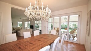 Ein geräumiger Essbereich mit einem großen Holztisch, weißen Stühlen, einer Chandelüster und Blick auf einen Garten durch Glastüren.