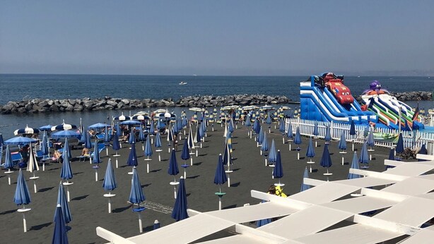 Ein Strand mit Reihen blauer Sonnenschirme und weißen Markisen, einer felsigen Küstenlinie und einer bunten Hüpfburg.