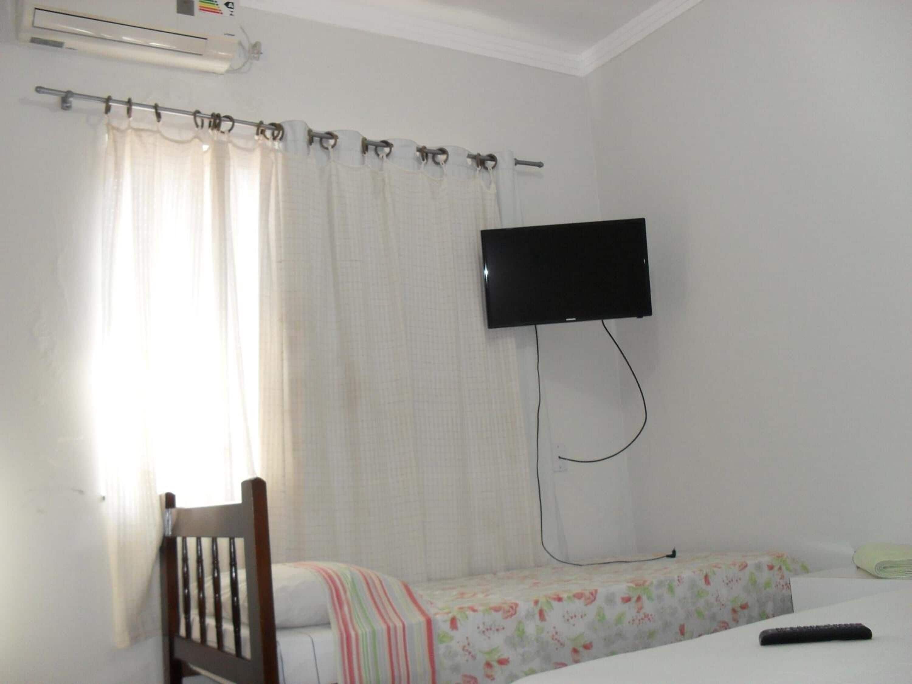 A single bed with a floral bedspread, a wooden chair, a television mounted on the wall, and a window with a curtain.
