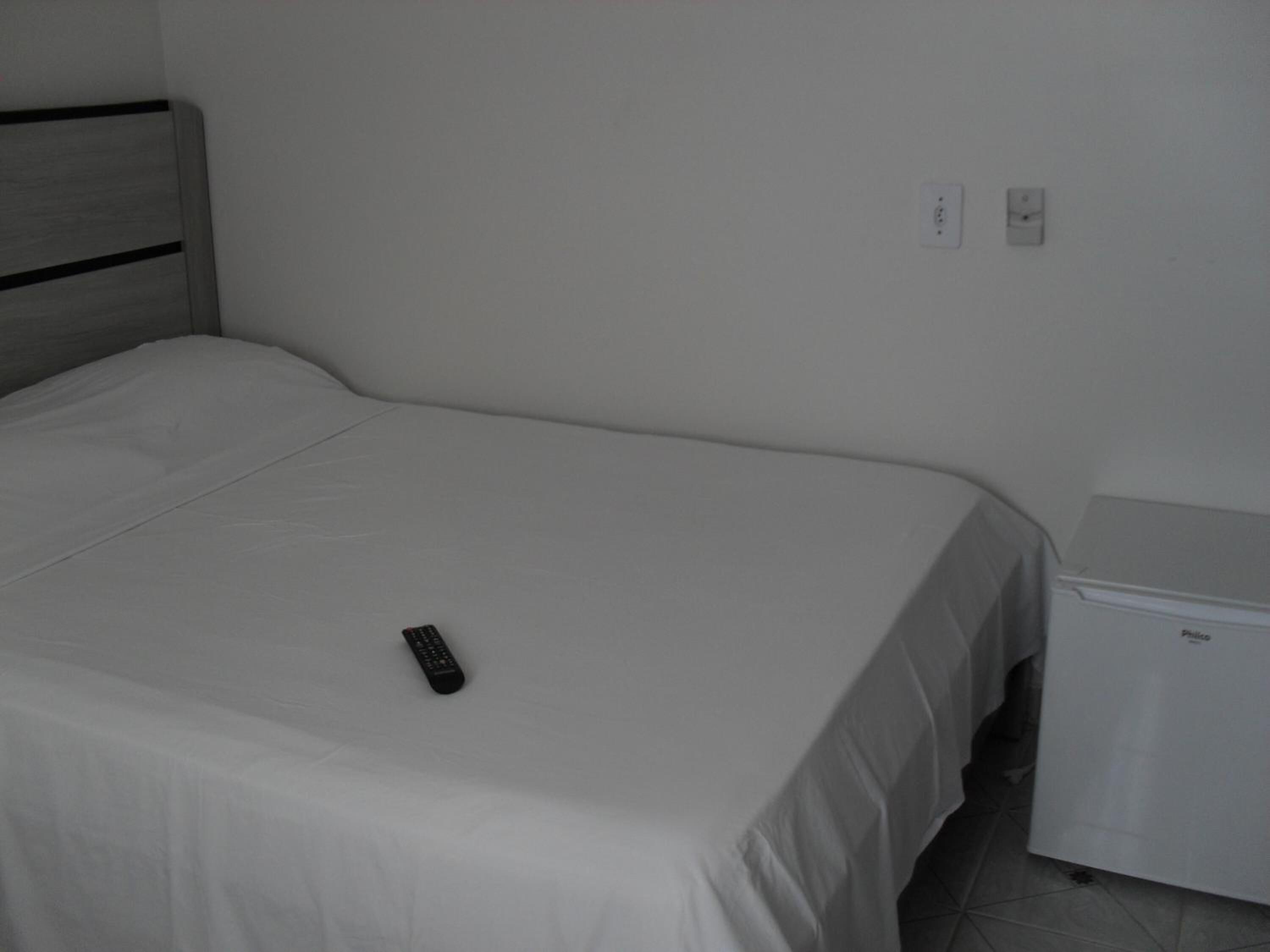 A single bed with a remote control on it, a small bedside table, and a wall socket.