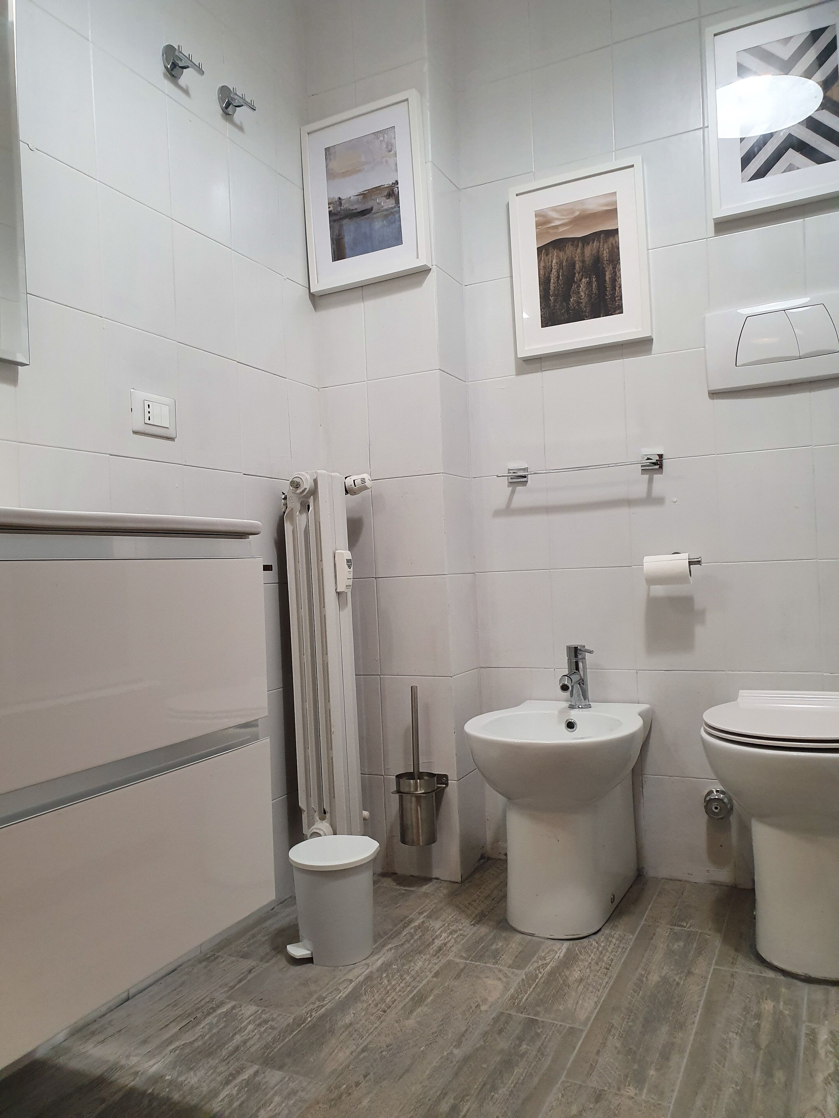 Une salle de bain moderne avec des carreaux blancs, un bidet, des toilettes, un lavabo et deux tableaux encadrés accrochés au mur.