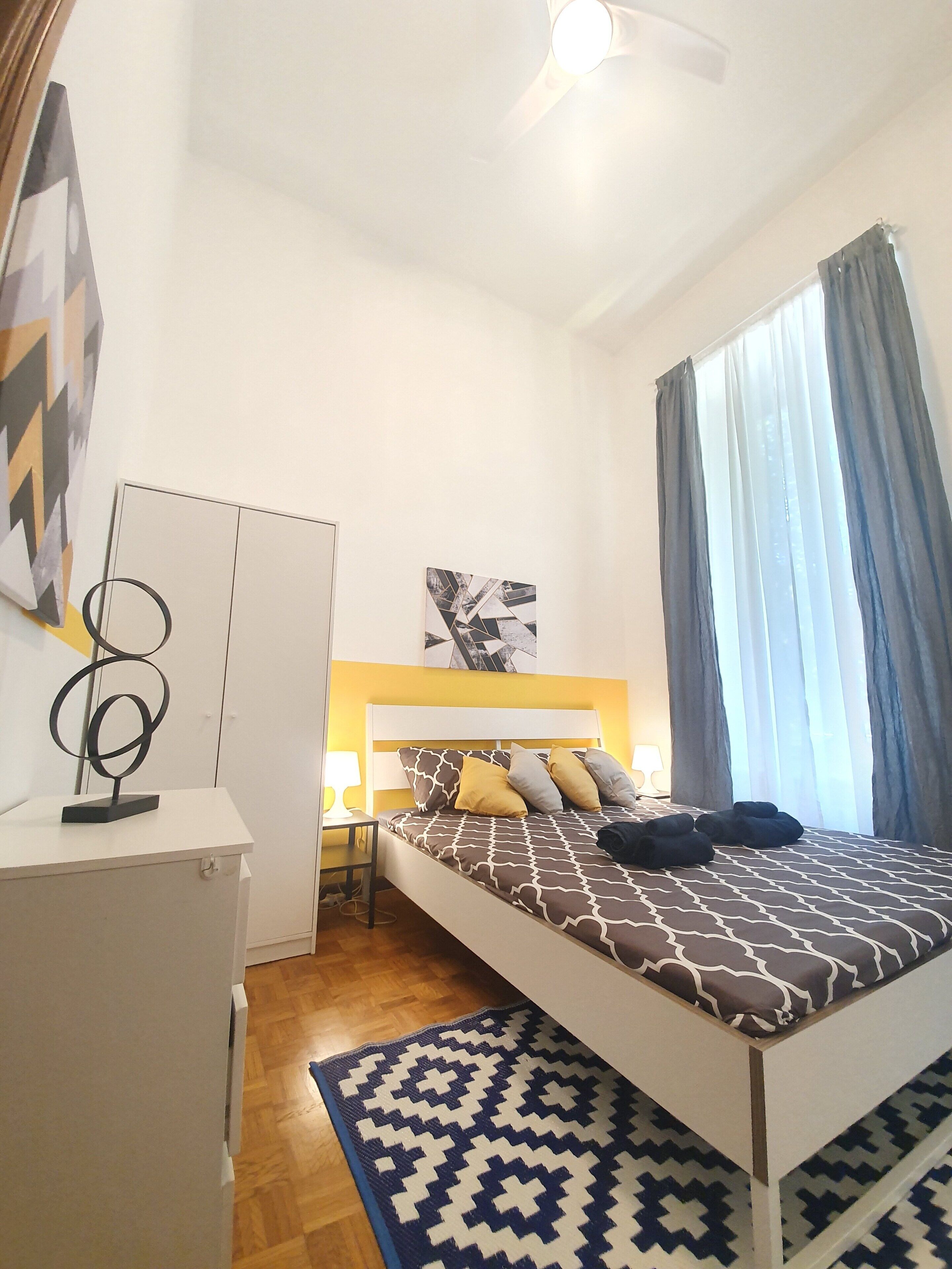 A modern bedroom with a bed, a desk, a chair, a lamp, and a window with curtains.