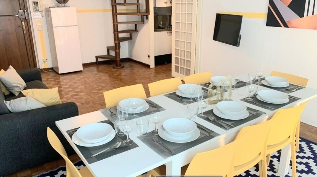 Zona pranzo con tavolo imbandito per quattro persone, sedie, divano, frigorifero, una scala e TV a parete.