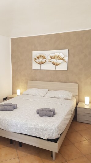 A neatly made bed with a patterned bedspread and two pillows, two rolled towels on the bed, a wall-mounted artwork of flowers, and two bedside lamps. - Casa Mudi (Turin)