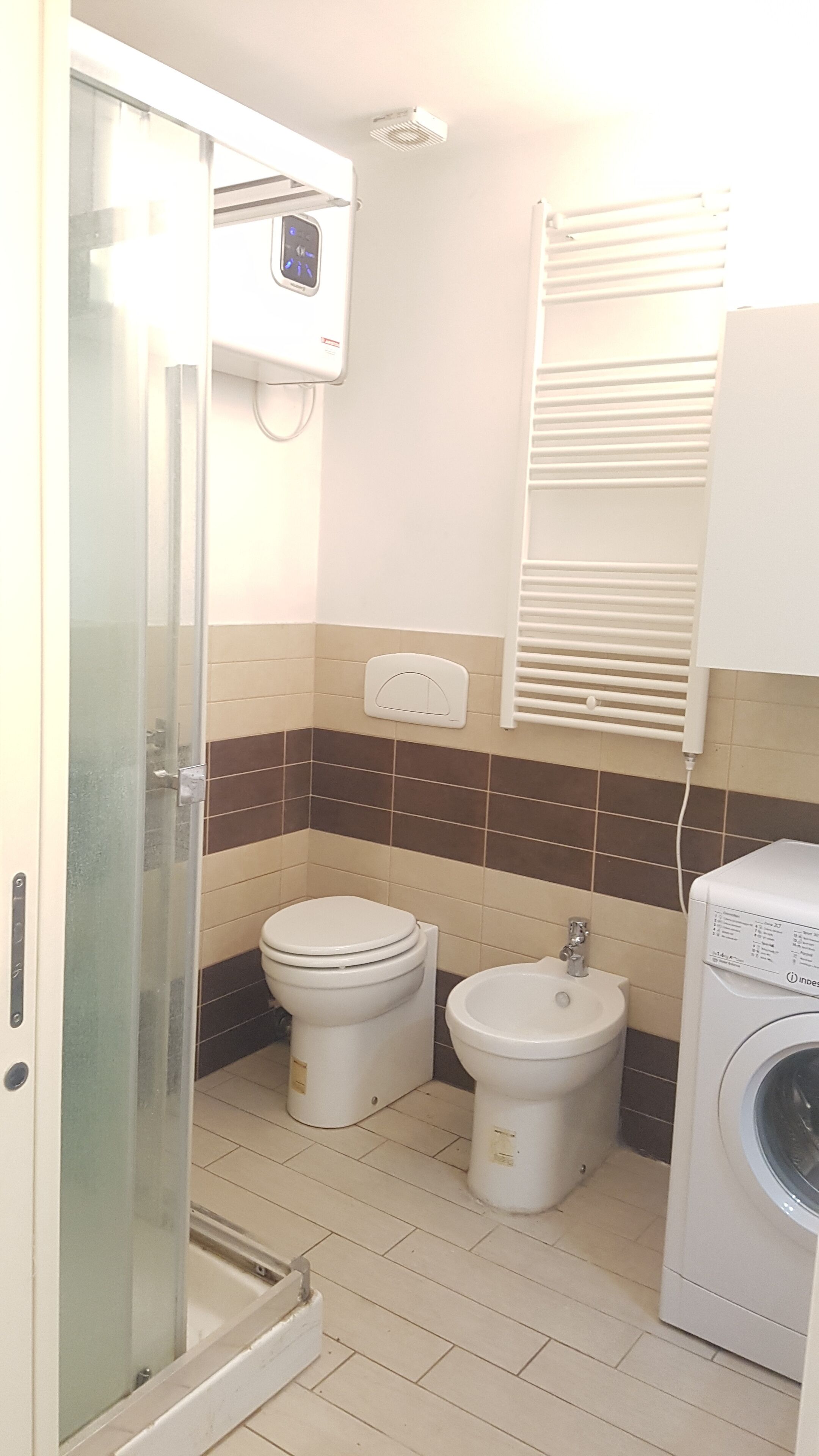 Casa de banho compacta com chuveiro envidraçado, sanita, bidé e máquina de lavar roupa.