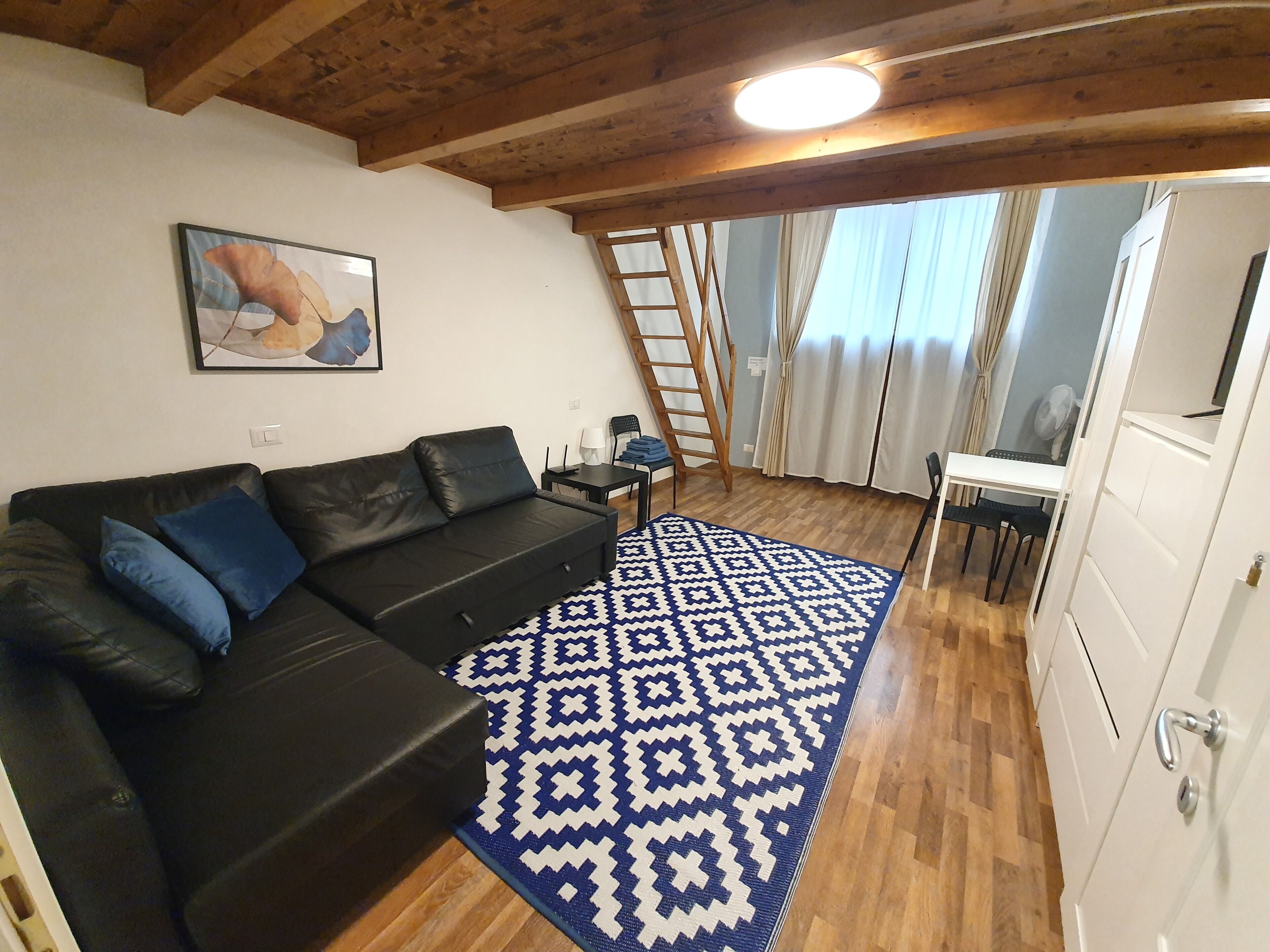 Una sala de estar acogedora con un sofá de cuero negro, una alfombra estampada, suelo de madera, un cuadro en la pared y una escalera en una esquina.