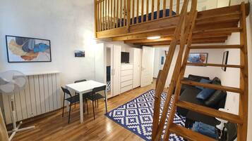 Un espace de type « loft » avec un escalier en bois, un coin repas comprenant une table et des chaises, et un coin salon avec un canapé et un tapis à motifs.