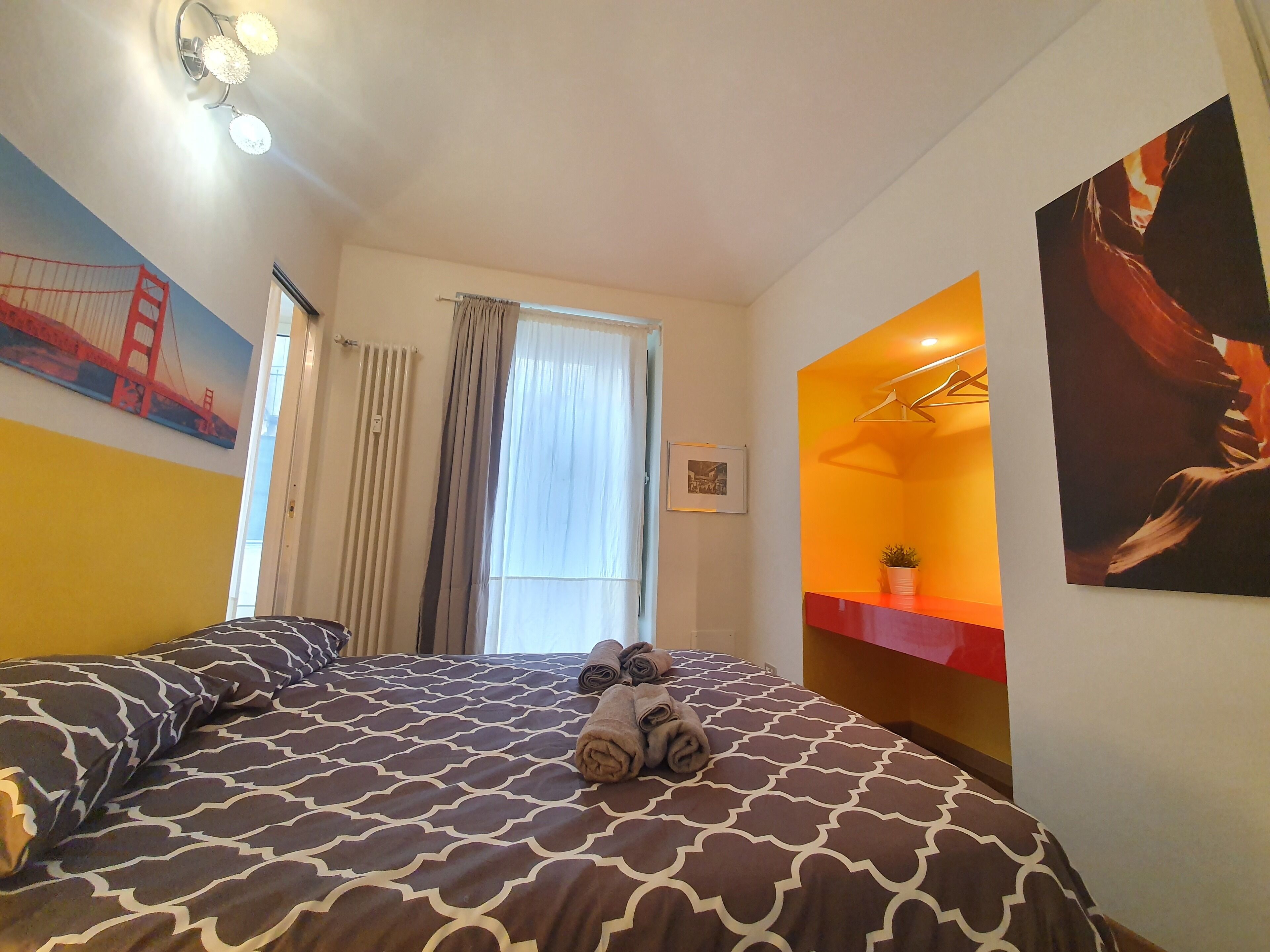 A hotel room with a bed, a window with curtains, a radiator, a wall with a picture of the Golden Gate Bridge, and a wall with a picture of a person.