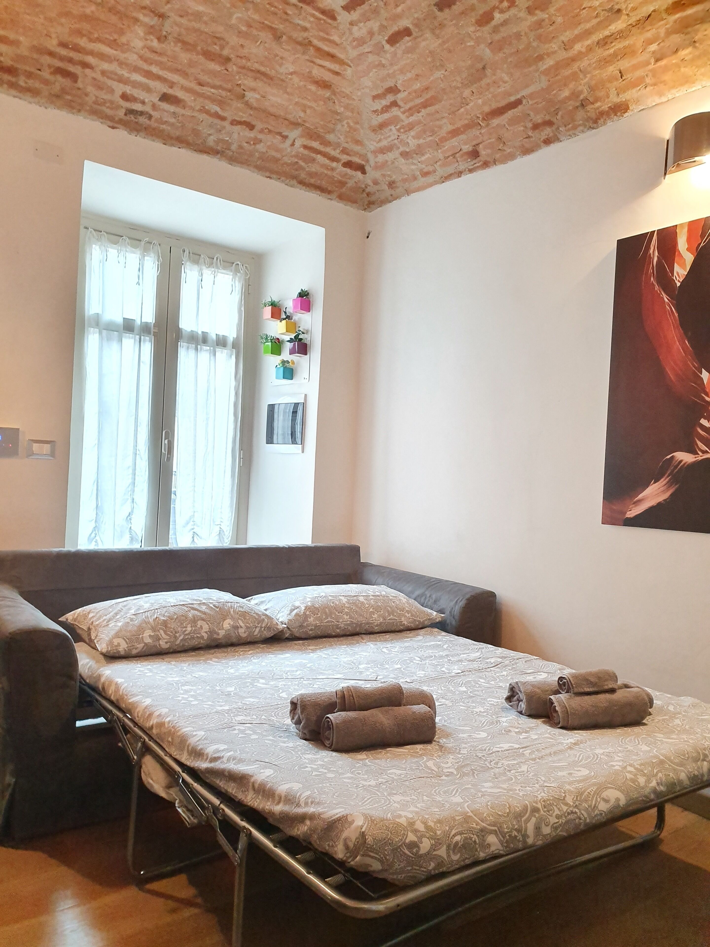 Ein Einzelbett mit gemustertem Bettbezug und zwei Kissen, ein Fenster mit Vorhängen, eine kleine Ablage mit Topfpflanzen und ein Bild an der Wand.