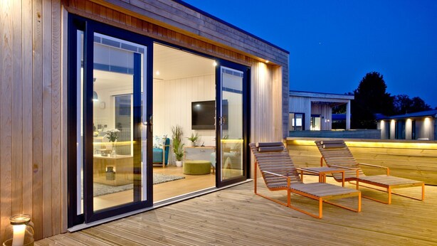Una casa moderna con amplios ventanales que dan a una sala de estar con televisor, asientos y una terraza de madera con sillones.