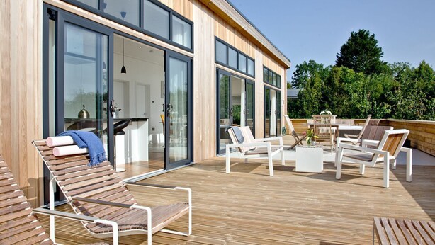 Una terraza moderna al aire libre con mobiliario de madera y puertas de vidrio que dan a una cocina.