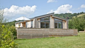 A modern wooden cabin with large windows and a deck, set in a grassy field with a hill in the background. - Perry Lodge, Strawberryfield Park - Hot Tub - Free Parking & WiFi (Cheddar)