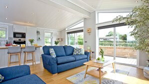 A modern living room with a blue sofa, wooden coffee table, and a kitchen area with a microwave and oven. - Perry Lodge, Strawberryfield Park - Hot Tub - Free Parking & WiFi (Cheddar)