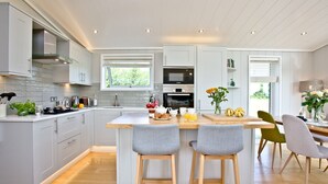 A modern kitchen with a central island, wooden stools, and a dining area with chairs. - Perry Lodge, Strawberryfield Park - Hot Tub - Free Parking & WiFi (Cheddar)