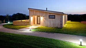 Une cabane en bois moderne, dotée d’une grande porte et d’une petite fenêtre, entourée d’une pelouse bien entretenue et d’un chemin de gravier menant à l’entrée.