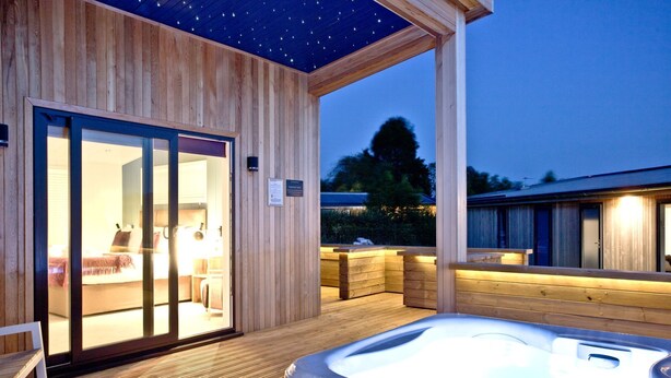 A wooden spa area with a hot tub, wooden deck, and a glass door leading to a room with a bed and seating area. - Batcombe Hollow, Strawberryfield Park (Cheddar)