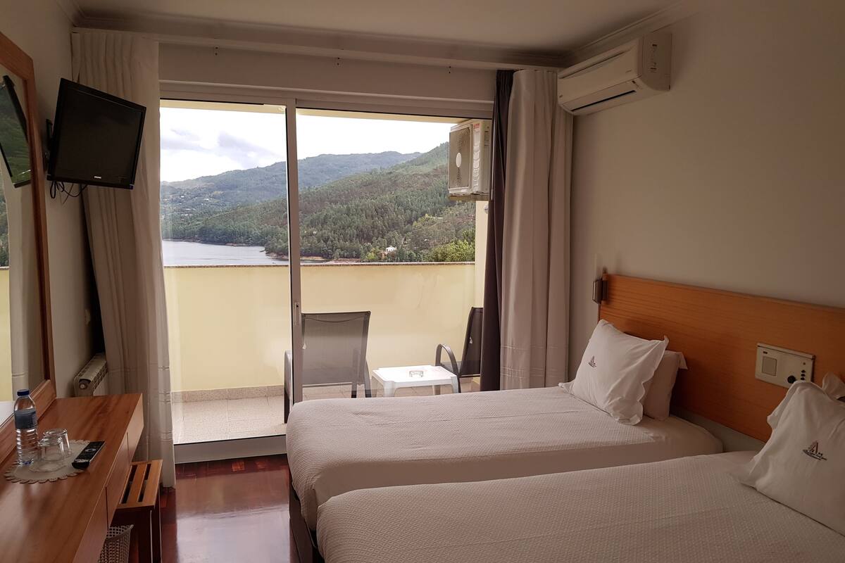 A hotel room with two beds, a TV, a table with a bottle and glasses, and a view of a mountainous landscape.