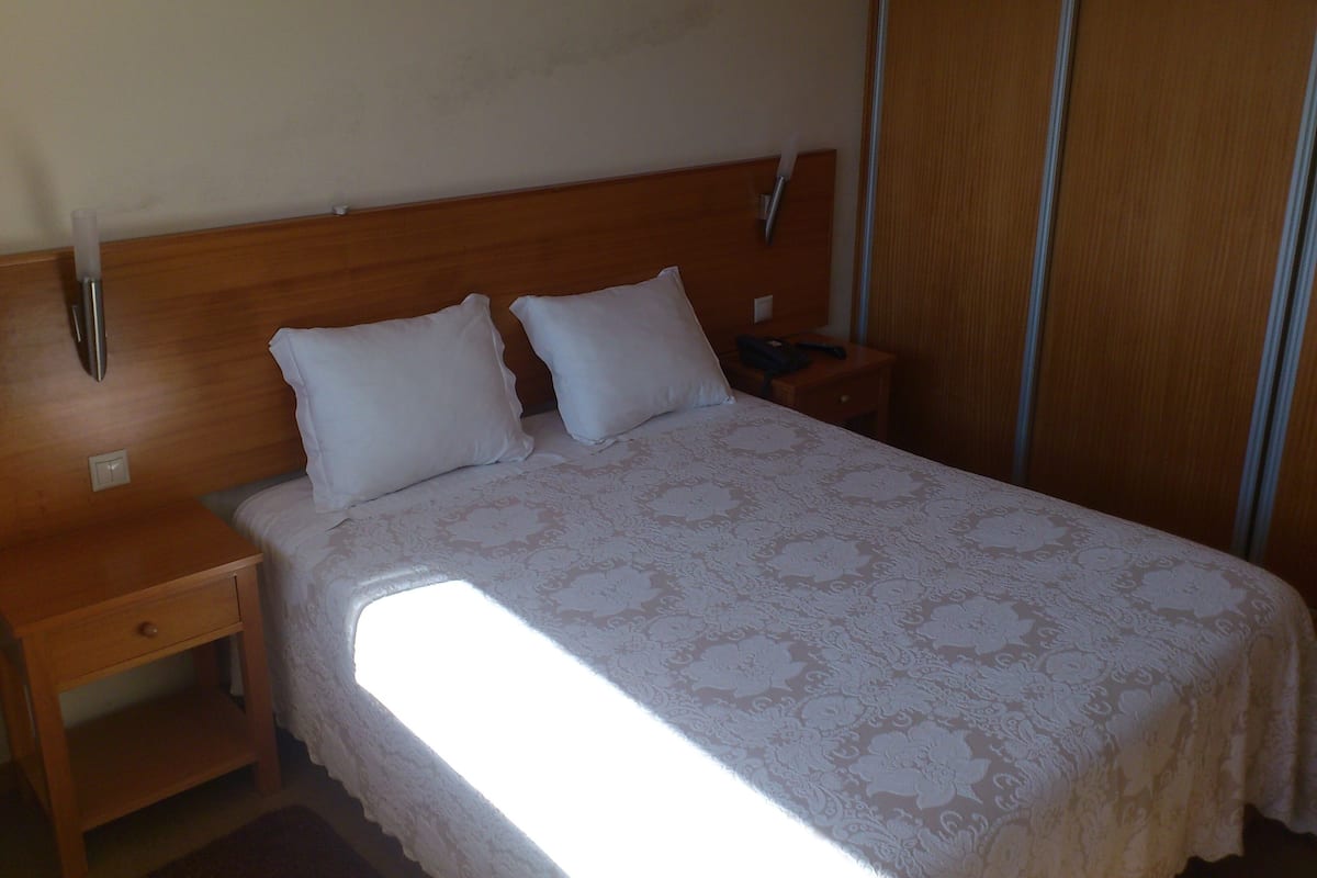 A hotel room with a wooden headboard, two white pillows, a bedside table with a lamp, and a large window providing natural light.