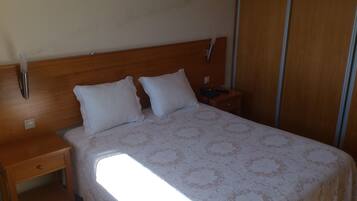 A hotel room with a wooden headboard, two white pillows, a bedside table with a lamp, and a large window providing natural light.