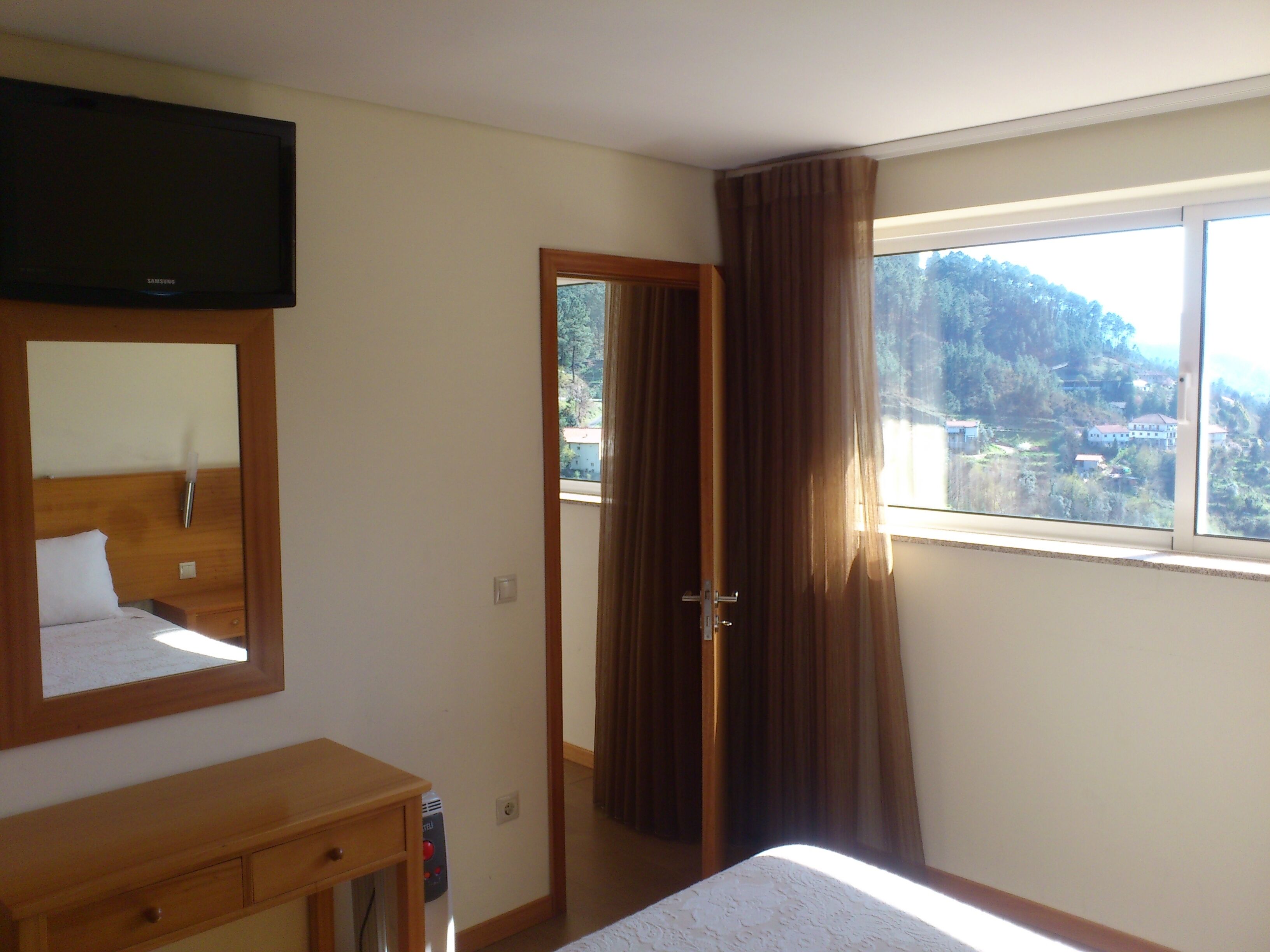 A hotel room with a bed, a wooden dresser, a television, a window with curtains, and a view of a mountainous landscape.