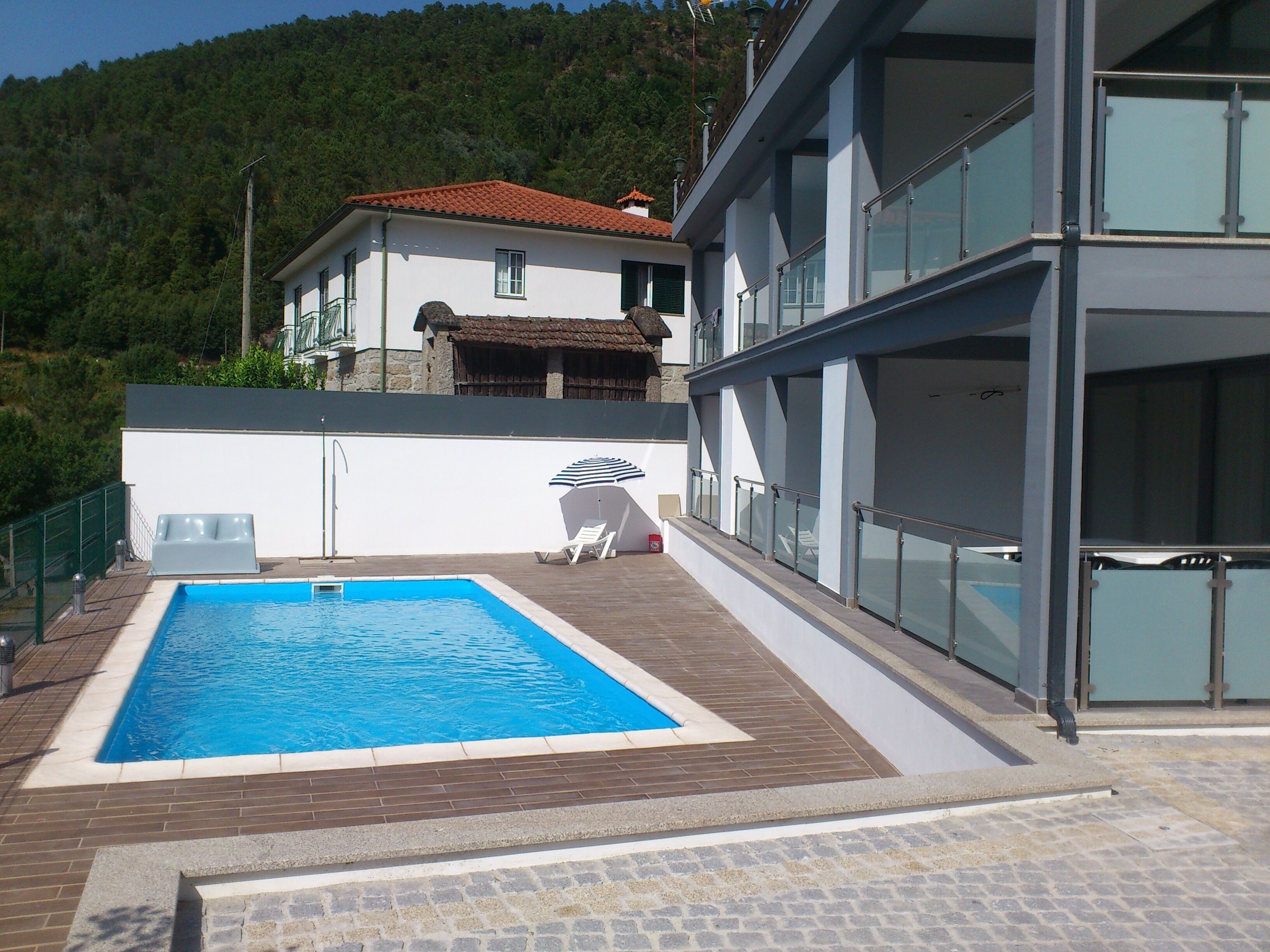 A swimming pool with a wooden deck, a white building with a red roof, and a glass railing.