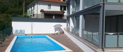 A swimming pool with a wooden deck, a white building with a red roof, and a glass railing.
